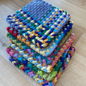 May include: A stack of six colorful woven potholders. Each potholder features a square shape with a braided edge. The colors include blue, green, yellow, purple, and orange. The potholders are stacked on top of each other.