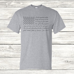 May include: A grey t-shirt featuring a black and white image of the American flag and the text "A well regulated militia being necessary to the security of a free state. the right of the people to keep and bear arms shall not be infringed. The Second Amendment to the Constitution of the United States of America. Dec. 1791"
