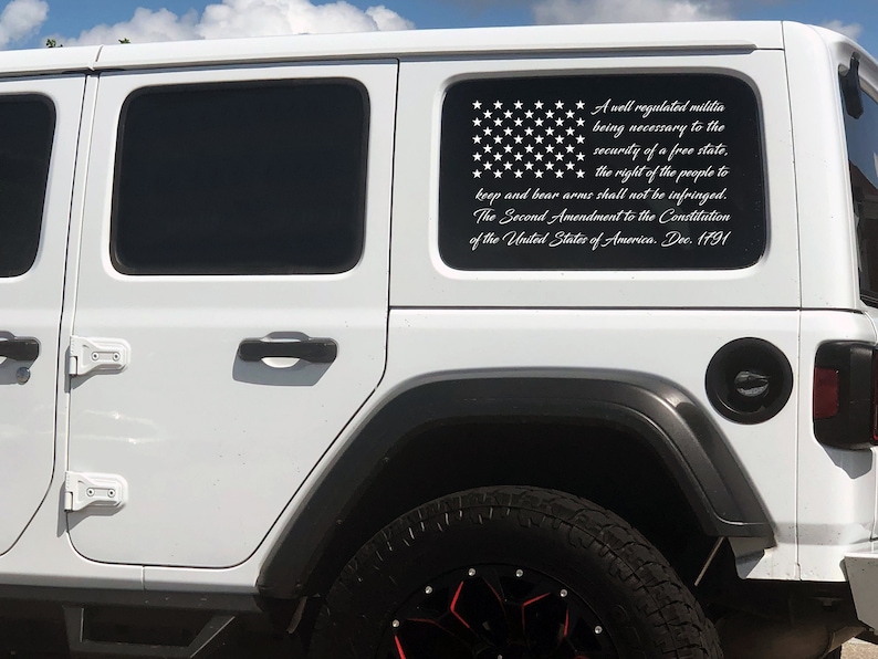 May include: A white Jeep with a black and white American flag decal on the rear window. The decal includes the text of the Second Amendment to the United States Constitution.