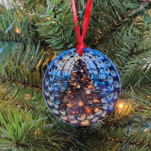 May include: Round Christmas ornament with a festive design. The ornament features a Christmas tree with lights inside a stained-glass window design. It hangs from a red ribbon against a backdrop of green Christmas tree branches.