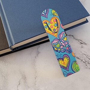 May include: A colorful bookmark with a heart design rests on top of two books. The bookmark features a blue background with yellow, pink, and green heart shapes. The books have blue and brown covers.