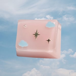 May include: A pink keycap with a light blue cloud design and gold star accents. The keycap is floating in front of a blue sky with white clouds.