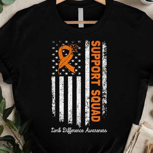 Puede incluir: Camiseta negra con un diseño de bandera estadounidense desgastada y una cinta naranja de concienciación. Las palabras "SUPPORT SQUAD" y "Limb Difference Awareness" están impresas en la camiseta. La camiseta se muestra en una percha de madera.