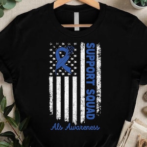 Peut inclure: T-shirt noir avec un motif de drapeau américain vieilli, un ruban bleu et les mots "SUPPORT SQUAD" et "Als Awareness" en bleu. Le t-shirt est présenté sur un cintre en bois.