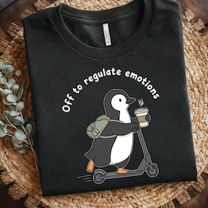 May include: Black t-shirt featuring a cartoon penguin riding a scooter and holding a coffee cup. The text "Off to regulate emotions" is printed above the penguin. The shirt is displayed on a woven basket.