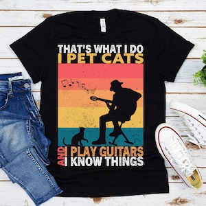 May include: Black t-shirt with a vintage-style graphic. The graphic features a silhouette of a person playing a guitar with a cat sitting in front of them. The text on the shirt reads "That's what I do I pet cats and I play guitars I know things."