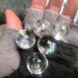 Op de afbeelding: Vijf heldere kwartskristallen bollen met iriserende regenboogkleuren. De bollen bevinden zich in de hand van een persoon die een grijze gebreide handschoen draagt.