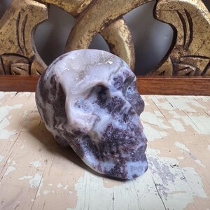 May include: A carved skull sculpture in shades of purple, white, and gray. The skull is made of a stone-like material and has detailed features. The sculpture is displayed on a distressed, light-colored wooden surface.