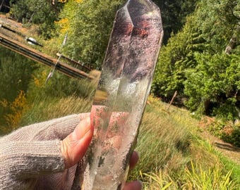 May include: A clear, elongated quartz crystal point with internal reddish-brown mineral inclusions. The crystal has a hexagonal, faceted structure with a pointed termination, displaying varying levels of transparency and natural internal textures.