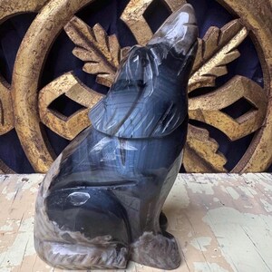 Puede incluir: Una escultura de lobo de piedra tallada en pose de aullido. El lobo es principalmente gris oscuro con acentos grises claros y marrones. La escultura se encuentra sobre una superficie de madera envejecida, con un fondo decorativo dorado.