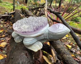 May include: A carved stone turtle sculpture with a large amethyst crystal shell. The turtle is gray and white with detailed flipper and head features. The amethyst crystal is a mix of clear and purple hues. The sculpture is displayed outdoors on a log.