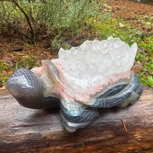 Peut inclure: Une sculpture de tortue en pierre sculptée avec une formation de cristaux de quartz clair sur son dos. Le corps de la tortue est gris, avec des touches de rose et de bleu. La sculpture est posée sur une surface en bois.