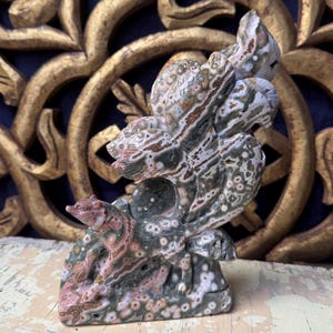 Può includere: Una scultura in pietra scolpita raffigurante un orso e altre figure di animali. La scultura è principalmente verde, rosa e marrone con un motivo maculato. L'opera d'arte è esposta su uno sfondo dorato e ornato.