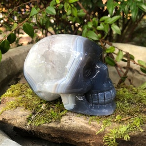 May include: A white and gray agate skull sculpture. The skull is carved from a single piece of agate and has a natural, organic look. The agate has a banded pattern of white, gray, and black.