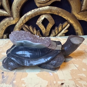 Peut inclure: Une figurine de tortue en pierre sculptée gris foncé avec une formation de cristal d'améthyste violet clair sur le dos. La tortue présente des marques de carapace détaillées et une finition lisse et polie. C'est un objet décoratif.