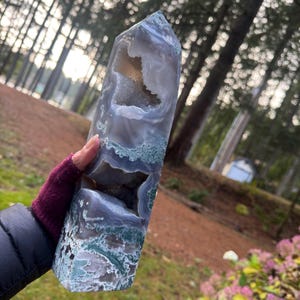 May include: A tall, polished agate crystal tower with a geode cavity. The stone features a mix of gray, blue, and green hues, with a natural, textured appearance. The crystal is held in a hand, with a blurred forest background.