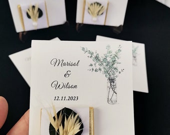May include: Wedding favor boxes with a white background and gold trim. The boxes feature a small bouquet of dried flowers and a pearl. The boxes are decorated with a watercolor illustration of eucalyptus leaves and the text "Marisol & Wilson 12.11.2023".