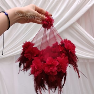 Puede incluir: Un ramo de tul y plumas burdeos con flores de tela roja. El ramo se sostiene en una mano con una pulsera azul de ojo turco.