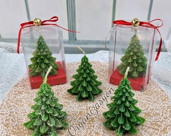May include: Green Christmas tree-shaped candles, some with glitter, are displayed. Two are in clear boxes with red bases and ribbon. The candles are on a decorative surface. The text "Kizce Organizasyon" is visible.