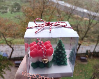 Könnte beinhalten: Eine transparente Geschenkbox enthält ein Arrangement aus roten, weißen und grünen Kerzen mit einer kleinen goldenen Glocke und einer grünen Weihnachtsbaumkerze. Die Box ist mit rot-weißem Bindfaden gebunden. Der Text "KizçeOrganizasyon" ist sichtbar.