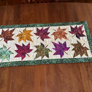 May include: A rectangular quilted table runner featuring ten appliqued fabric maple leaves in various colors including red, orange, yellow, purple, and green. The leaves are set against a cream background with swirl quilting and a green border.