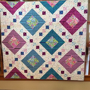 May include: A white quilt with a diamond pattern. The diamonds are made of different fabrics with stripes, floral patterns, and polka dots. The quilt is made with pink, blue, and brown fabrics.