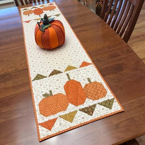 May include: A quilted fabric table runner featuring a cream background with a small floral print and appliqued orange pumpkin motifs. A stuffed fabric pumpkin with orange segments and a dark green stem sits on the runner.