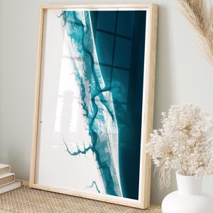 May include: A framed abstract art print depicting a coastal scene in teal and white. The artwork is displayed on a light wooden bench with woven detailing, alongside a white vase with dried flowers and decorative pampas grass.