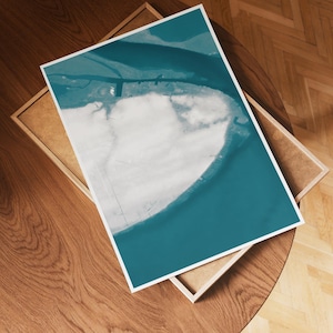 May include: A framed print depicting an aerial view of a coastal landscape. The artwork shows a white landmass surrounded by teal waters and a teal sky. The print is set in a wooden frame, resting on a wooden table.