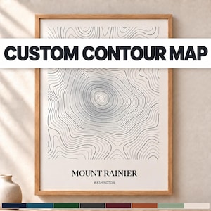 May include: A framed wall art print featuring a black line contour map of Mount Rainier, Washington, on a cream background. The text CUSTOM CONTOUR MAP is printed in bold black letters across the center of the rectangular wooden frame.