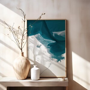 May include: A framed abstract art print with shades of teal and white, resembling an aerial view of a coastline. A light-colored wooden vase holds branches with small buds. A small white vase and a decorative ceramic piece are also on display.