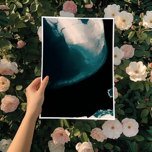 Op de afbeelding: Een abstracte print met tinten van teal en wit tegen een zwarte achtergrond. Het kunstwerk wordt getoond tegen een achtergrond van weelderig groen gebladerte en zachte roze en witte rozen.