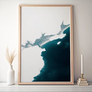 May include: Abstract art print featuring a coastal landscape with a contrast of white and deep teal. The artwork is framed in a light wood frame. A white vase with dried pampas grass and a candle on a stack of books are in the foreground.