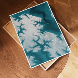 May include: A framed art print featuring an abstract aerial view in shades of teal and white. The artwork is displayed on a wooden table, showcasing a detailed landscape design. The print is encased in a white border and a wooden frame.