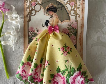 Op de afbeelding: Een handgemaakte wenskaart met een vintage illustratie van een vrouw met een paraplu. De kaart is versierd met een gele zakdoekjurk met roze rozen en groene bladeren. Een roze strik accentueert de jurk. De achtergrond heeft een bloemenpatroon.