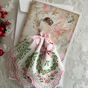 Op de afbeelding: Een kerstkaart met een engelillustratie, roze vleugels en een bloemenkrans. Een witte zakdoek met een roze geschulpte rand en een hulst- en bessenontwerp is bevestigd met een roze strik. De kaart ligt op een witte envelop.