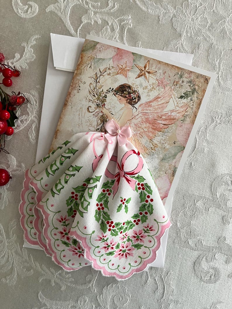 Op de afbeelding: Een kerstkaart met een engelillustratie, roze vleugels en een bloemenkrans. Een witte zakdoek met een roze geschulpte rand en een hulst- en bessenontwerp is bevestigd met een roze strik. De kaart ligt op een witte envelop.