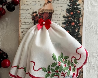 Op de afbeelding: Een kerstkaart met een vintage illustratie van een vrouw in een rode jurk en een witte zakdoekjurk met hulst- en belmotieven. Een rode strik siert de zakdoekjurk. Rode bessen bevinden zich aan de linkerkant.