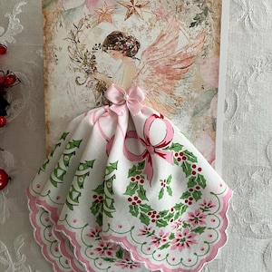 Op de afbeelding: Een kerstkaart met een engelillustratie met roze vleugels en een bloemenkrans. De jurk van de engel is een witte zakdoek met een roze strik, hulst en bloemenmotief. De kaart heeft een vintage uitstraling.