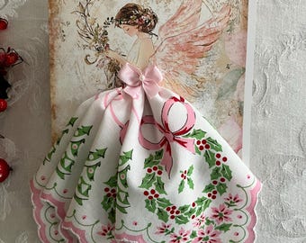 Op de afbeelding: Een kerstkaart met een engelillustratie met roze vleugels en een bloemenkrans. De jurk van de engel is een witte zakdoek met een roze strik, hulst en bloemenmotief. De kaart heeft een vintage uitstraling.