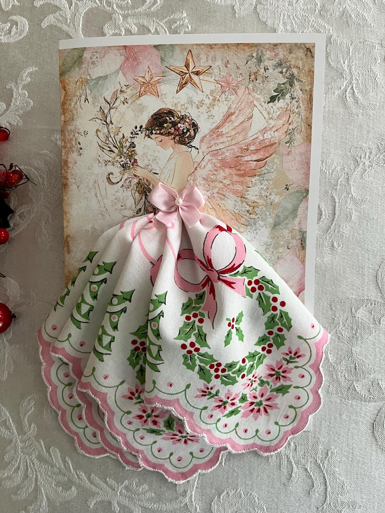 Op de afbeelding: Een kerstkaart met een engelillustratie met roze vleugels en een bloemenkrans. De jurk van de engel is een witte zakdoek met een roze strik, hulst en bloemenmotief. De kaart heeft een vintage uitstraling.