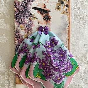 May include: A greeting card featuring a watercolor illustration of a woman in a hat, with a light blue handkerchief adorned with purple flowers and green leaves. The handkerchief has a pink border and a purple bow.