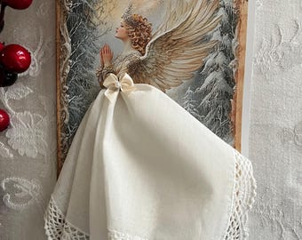 Op de afbeelding: Een wenskaart met een engel met grote vleugels, die een kroon draagt en bidt. Een witte zakdoek met een gehaakte kanten rand is met een strik aan de kaart bevestigd. De achtergrond toont een besneeuwde bosscène.