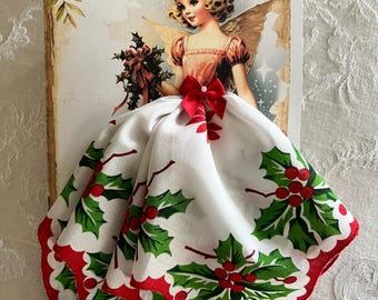 Op de afbeelding: Een vintage kerstkaart met een retro engel illustratie. De jurk van de engel is gemaakt van een witte zakdoek met een hulst- en bessenpatroon, afgewerkt met rode geschulpte randen. Een rode strik siert de taille.