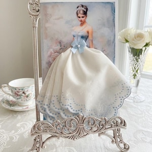 May include: A decorative silver metal picture frame with an ornate design holds a portrait of a woman in a blue and white gown. A white fabric with blue trim is draped over the portrait, resembling the dress. A teacup and vase of white roses are in the background.
