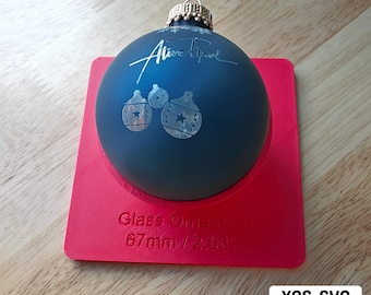 May include: A dark blue glass Christmas ornament with a white star design and the text "Alive Pixel" on the front. The ornament is 2.63 inches in diameter and is sitting on a red base.