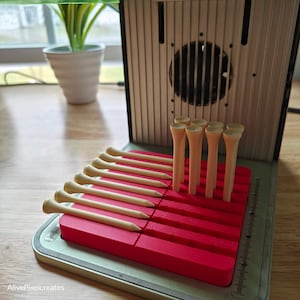 Op de afbeelding: Een rode golf tee houder met verschillende houten golf tees. De houder is rechthoekig en heeft sleuven om de tees vast te houden. De houder staat op een houten oppervlak. De tekst "AlivePixelcreates" is zichtbaar.
