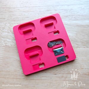 Könnte beinhalten: Eine leuchtend pinkfarbene Plastikform mit fünf rechteckigen Vertiefungen in verschiedenen Größen. Eine silberne Halskette mit einem kleinen silbernen Briefumschlag-Anhänger liegt in einer der Vertiefungen.