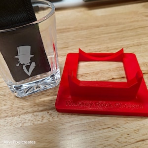 May include: A red 3D printed coaster with the text "Whiskey Rock Glasses" on the bottom. The coaster is designed to hold a glass of whiskey.