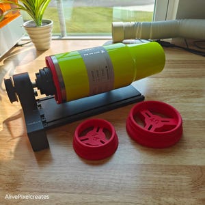 May include: A black and gray 3D printer with a yellow and red tumbler in the center. Two red plastic rings are on the table next to the printer. The larger ring has the text "4.0" printed on it. The smaller ring has the text "7.4" printed on it.